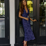 A woman in a deep purple floral dress leaning on the outside of a house holding a champagne glass.