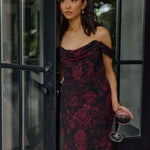 Woman in a dark floral dress holding a glass of red wine, standing in a doorway.
