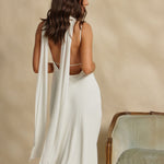 Woman in a white dress with a scarf draped around her neck standing against a beige wall.