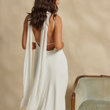 Woman in a white dress with a scarf draped around her neck standing against a beige wall.