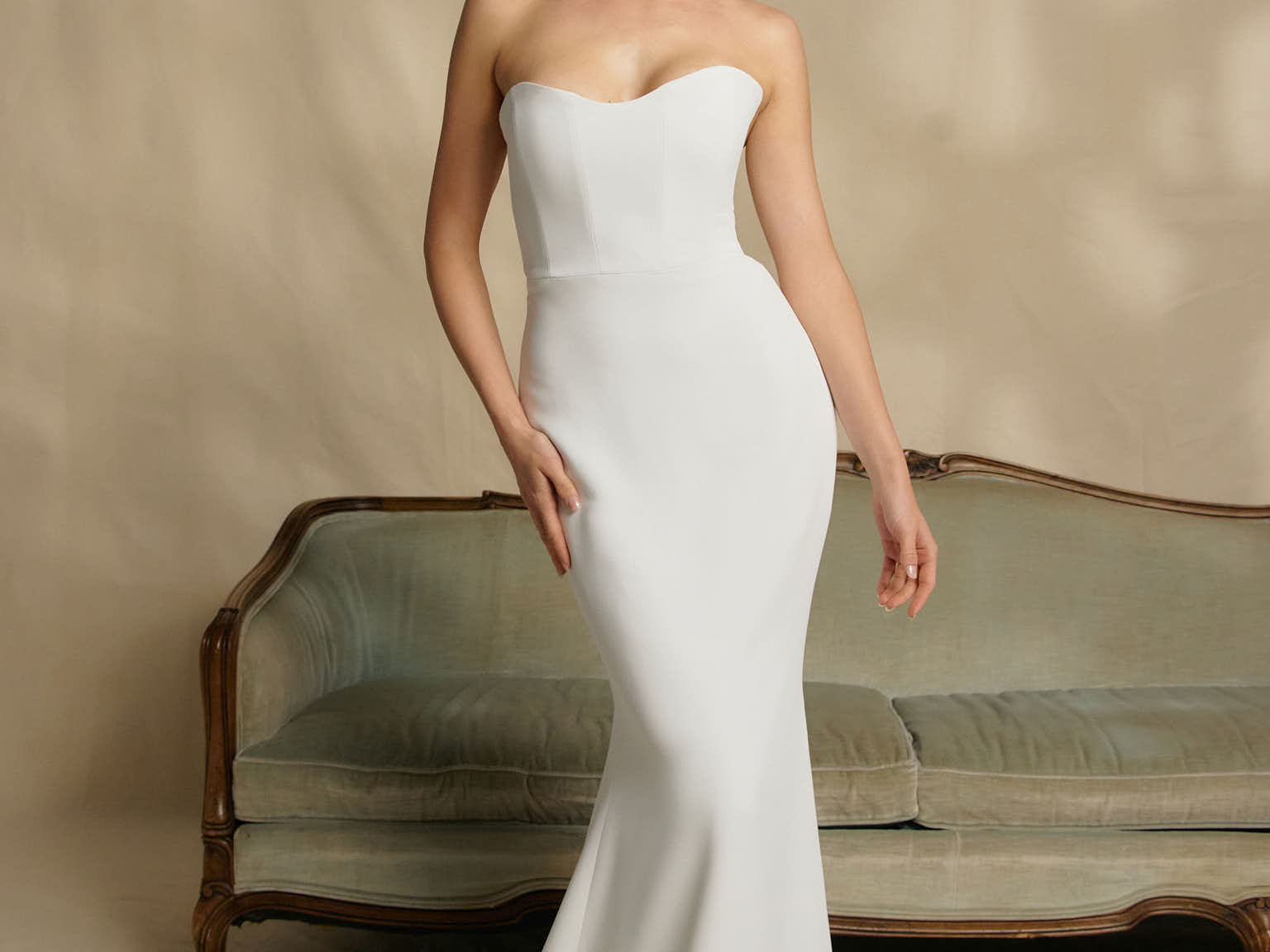 Woman in a strapless white wedding dress standing in front of a vintage couch.