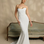 Woman in a strapless white wedding dress standing in front of a vintage couch.