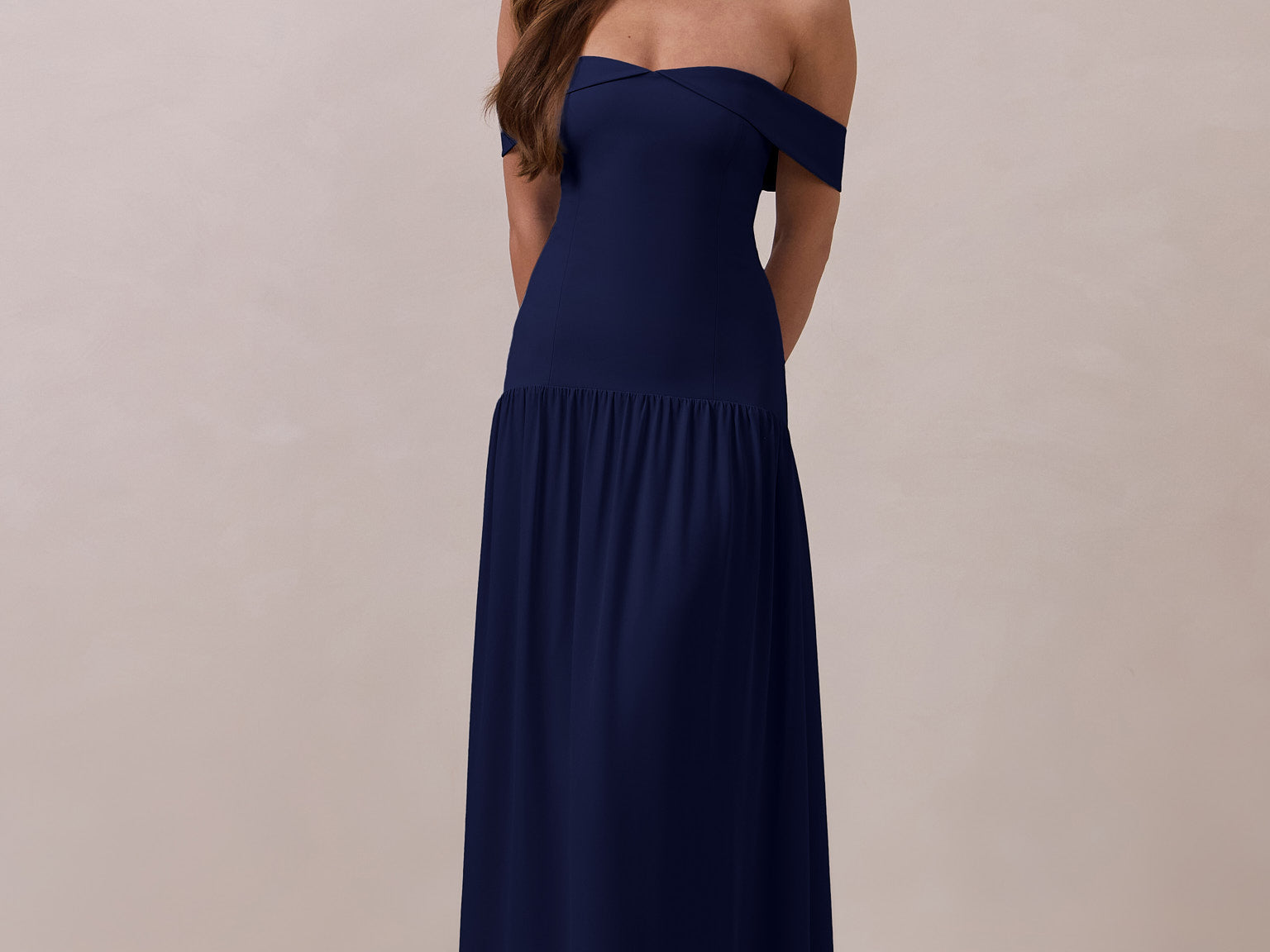 Woman wearing a navy off-shoulder gown with a drop waist standing against a beige background
