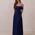 Woman wearing a navy off-shoulder gown with a drop waist standing against a beige background