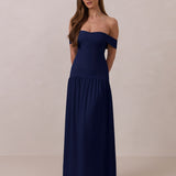 Woman wearing a navy off-shoulder gown with a drop waist standing against a beige background