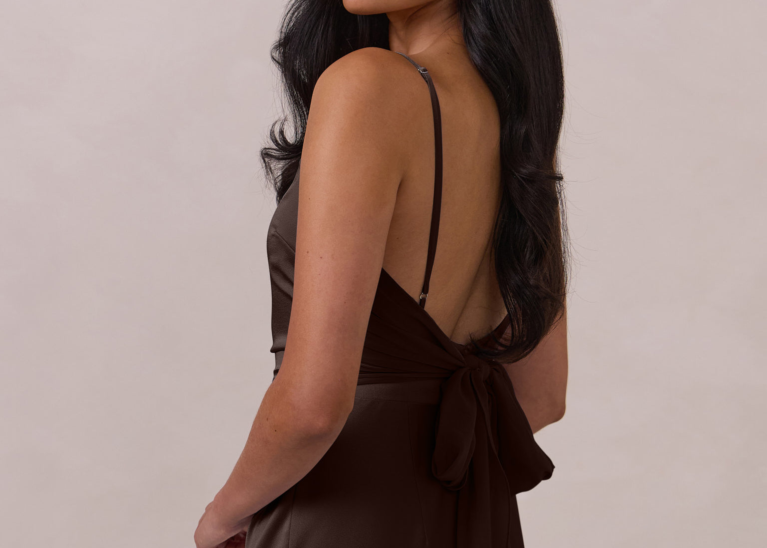 Woman wearing a brown coloured dress with thin straps and a tied back with bow detail on a plain background