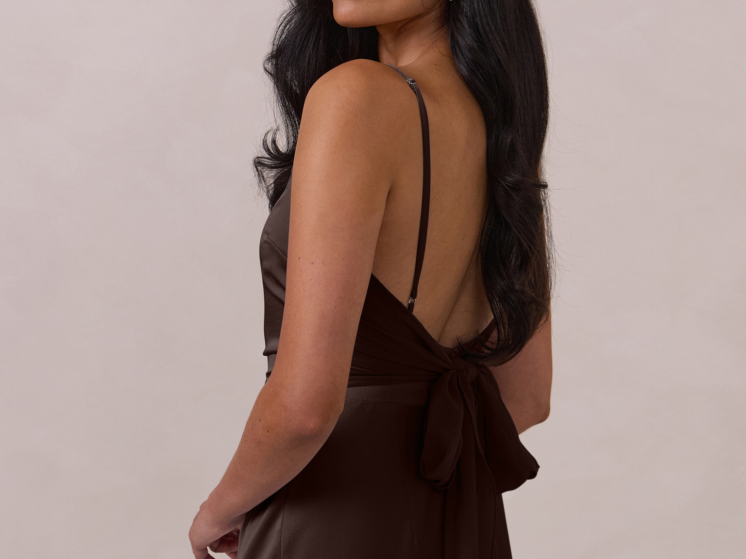 Woman wearing a brown coloured dress with thin straps and a tied back with bow detail on a plain background