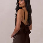 Woman wearing a brown coloured dress with thin straps and a tied back with bow detail on a plain background