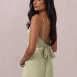 Woman wearing a pistachio coloured dress with thin straps and a tied back with bow detail on a plain background