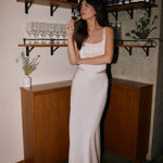 Woman in a off white satin set standing in a bar setting with wine bottles and glasses on shelves behind her