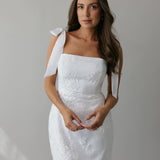 A woman stands against a light grey backdrop. She's wearing a white mini dress with white floral embroidery and satin tie straps. This bridal event dress is called the Tanner Mini.
