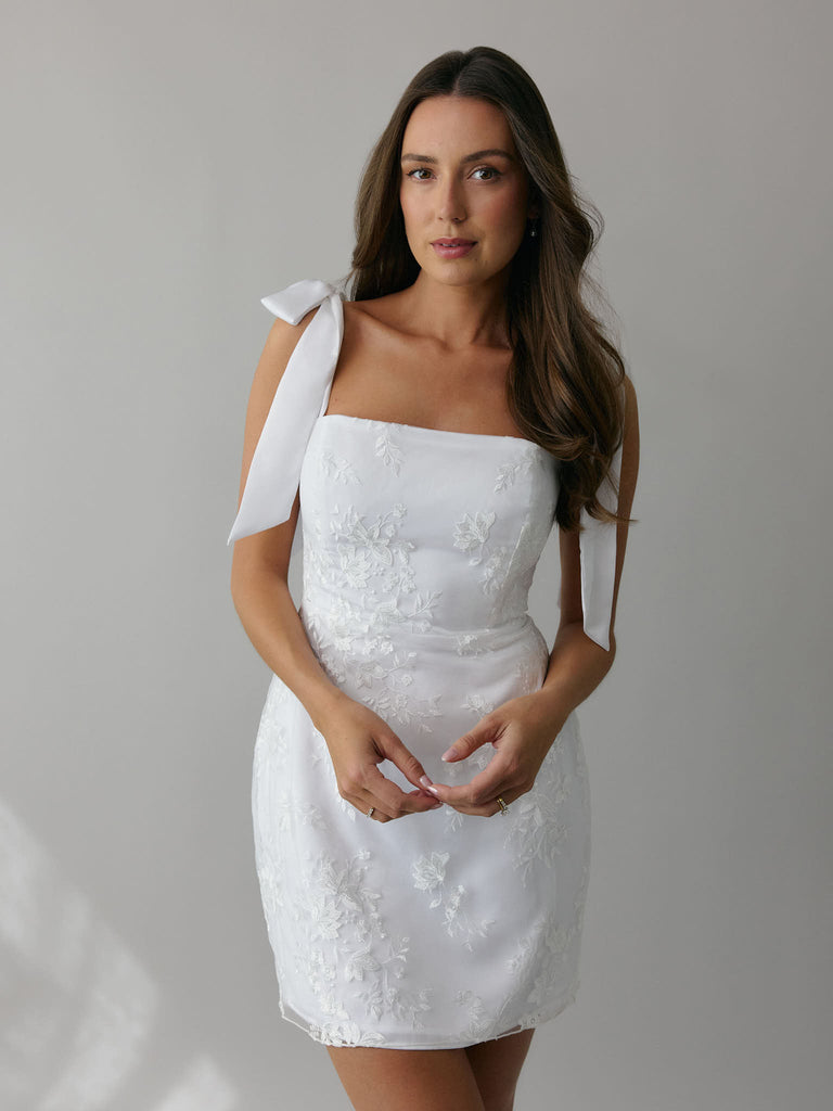 A woman stands against a light grey backdrop. She's wearing a white mini dress with white floral embroidery and satin tie straps. This bridal event dress is called the Tanner Mini.